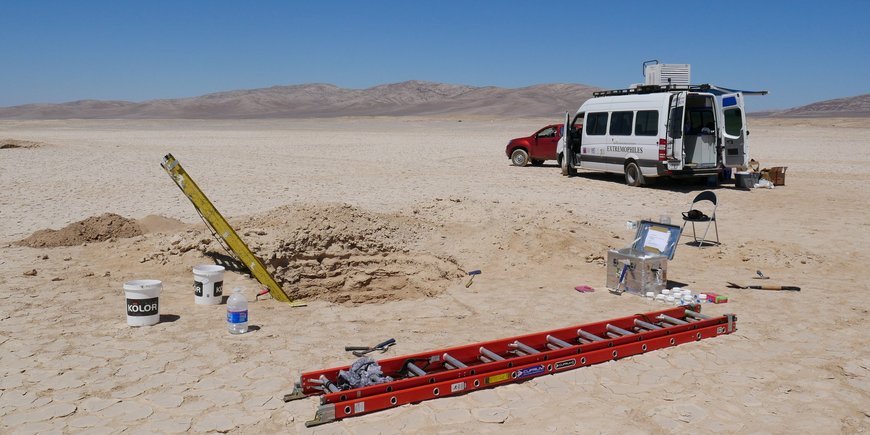 In the foreground an excavated pit with a yellow ladder. A red ladder lies on the ground. Further back is a white box van. All around: desert. Blue sky.